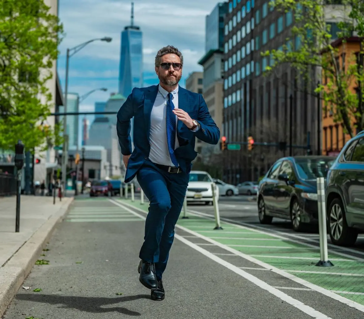 Hombre de negocios con traje azul y gafas de sol corriendo por la calle de la ciudad con rascacielos al fondo.