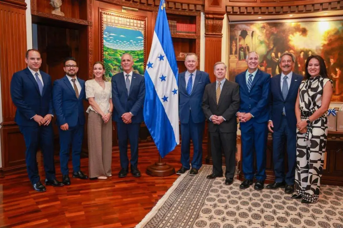 Grupo de personas en traje formal posando junto a una bandera nacional en una oficina ejecutiva.