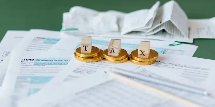 Primer plano de formularios de impuestos, incluyendo el 1040, con tres pilas de monedas doradas y bloques de madera que forman 'TAX'. Un bolígrafo plateado y documentos doblados completan la escena financiera.