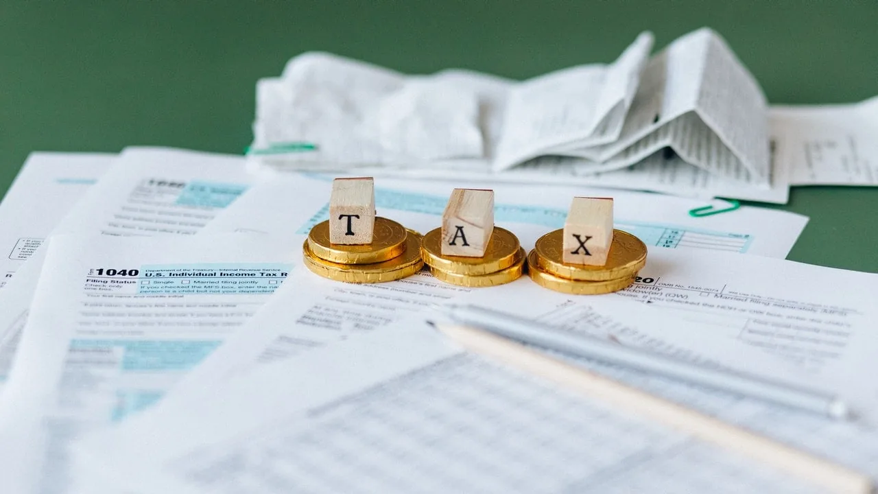 Primer plano de formularios de impuestos, incluyendo el 1040, con tres pilas de monedas doradas y bloques de madera que forman 'TAX'. Un bolígrafo plateado y documentos doblados completan la escena financiera.