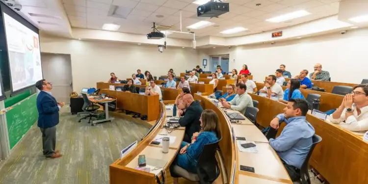 Un orador presenta en un aula moderna con asientos escalonados a un grupo diverso de profesionales atentos. La pantalla muestra una diapositiva.