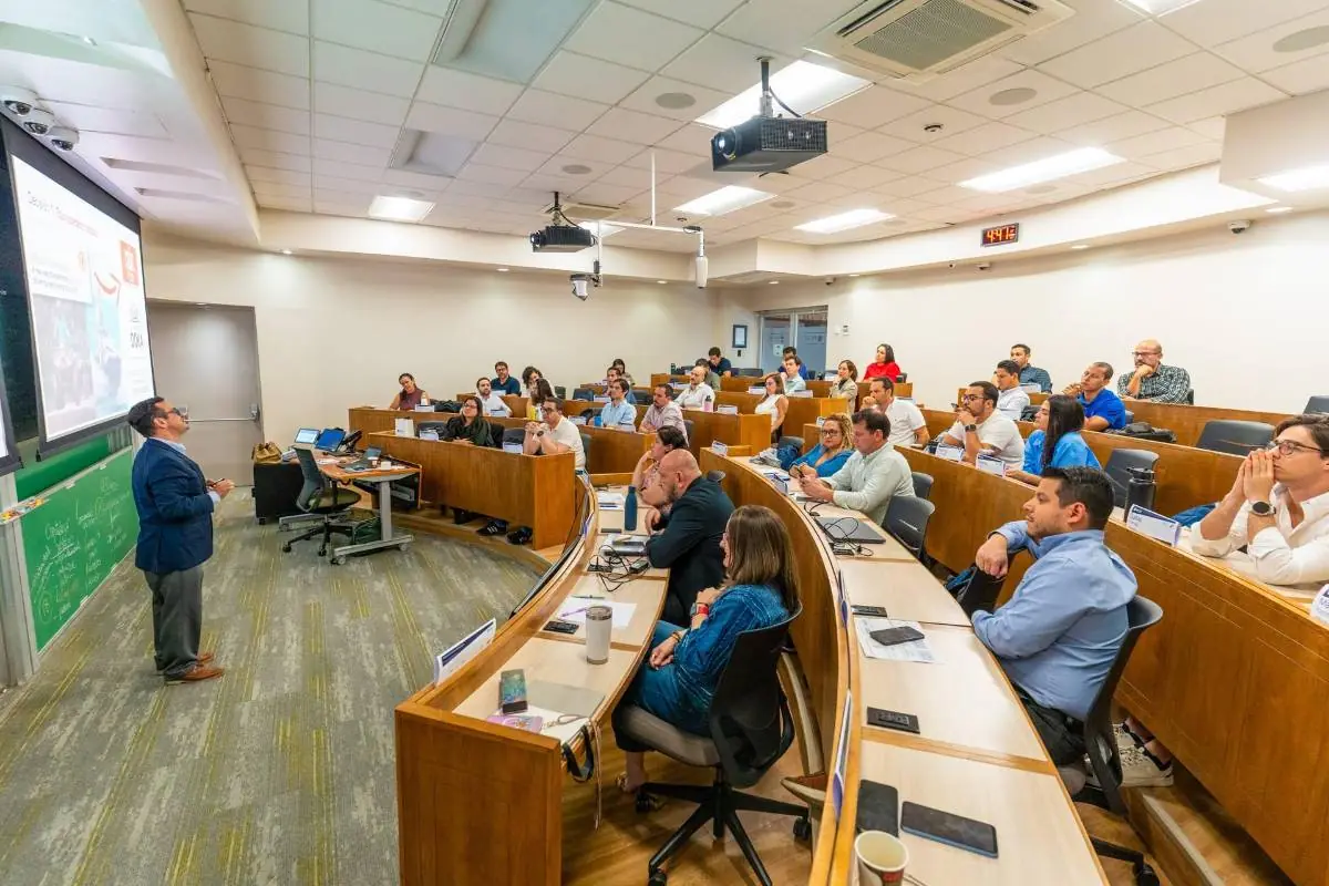 Un orador presenta en un aula moderna con asientos escalonados a un grupo diverso de profesionales atentos. La pantalla muestra una diapositiva.