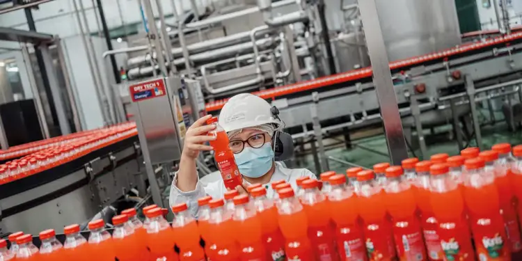 Trabajadora con casco y mascarilla inspecciona una botella de bebida en una línea de embotellado industrial.