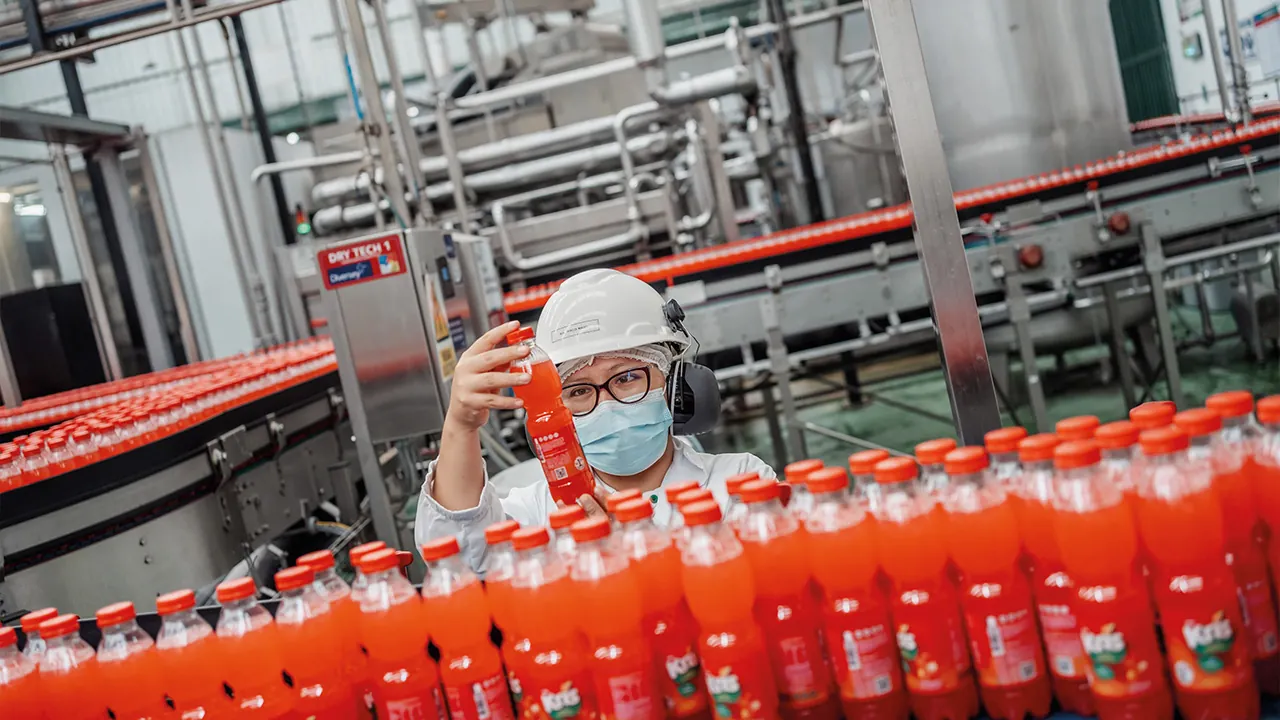Trabajadora con casco y mascarilla inspecciona una botella de bebida en una línea de embotellado industrial.