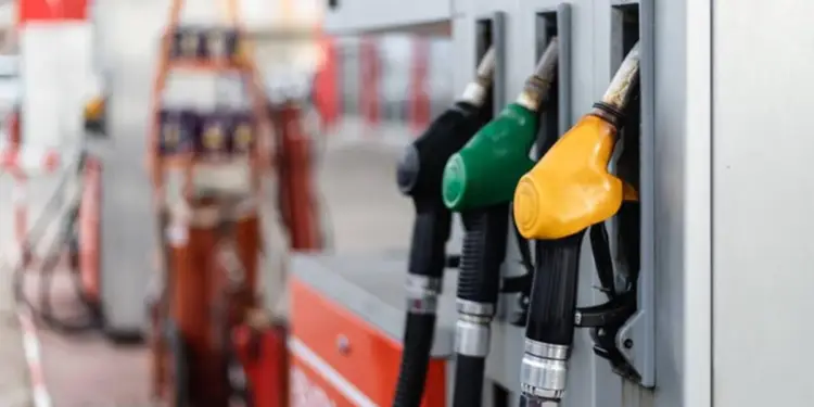 Tres boquillas de gasolina de colores (negro, verde, amarillo) colgando de una bomba en una estación de servicio, representando el costo del combustible.