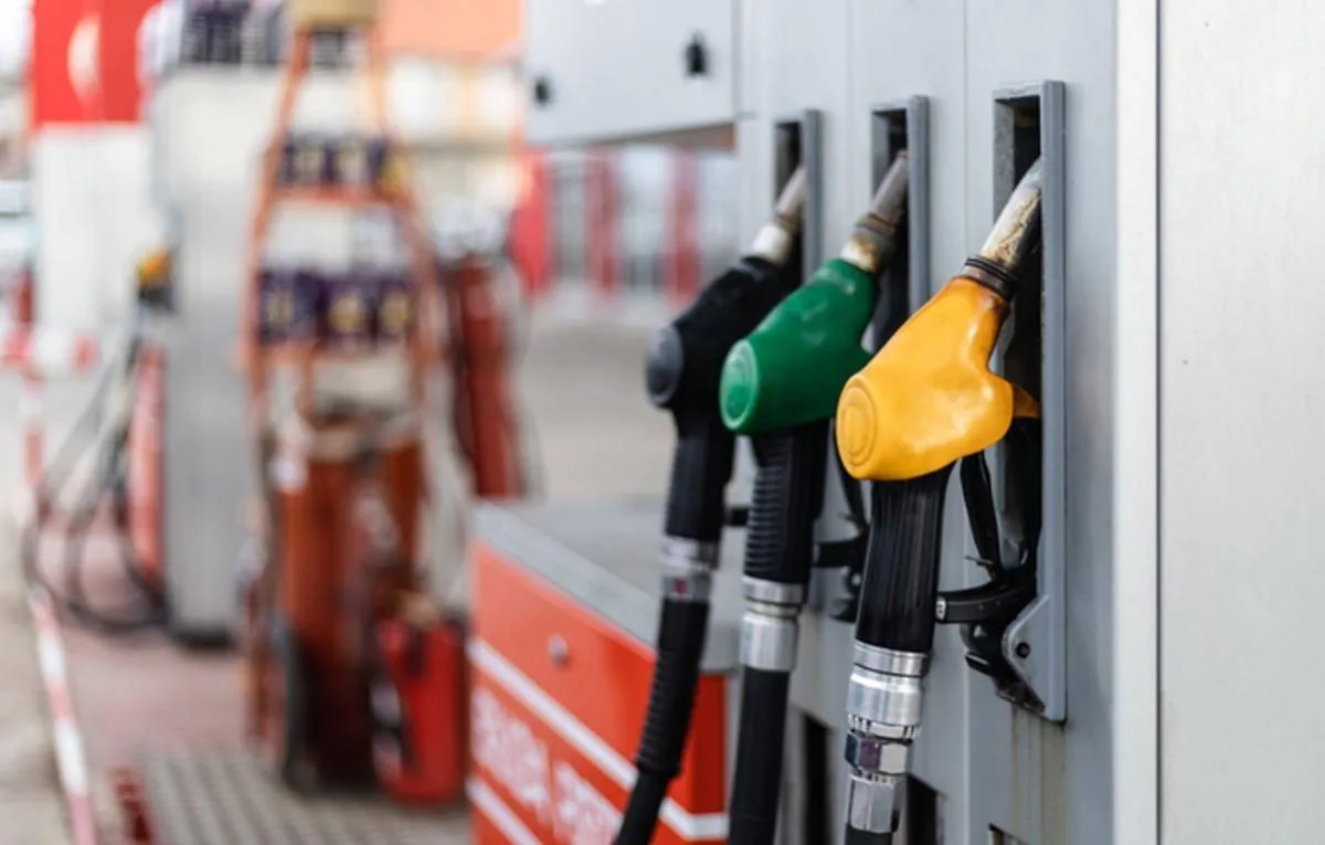 Tres boquillas de gasolina de colores (negro, verde, amarillo) colgando de una bomba en una estación de servicio, representando el costo del combustible.