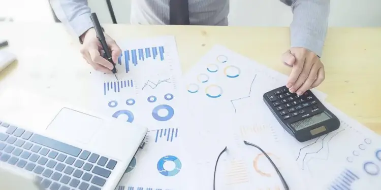 Persona analizando documentos financieros con gráficos y datos, usando una calculadora y un portátil en un escritorio de madera.