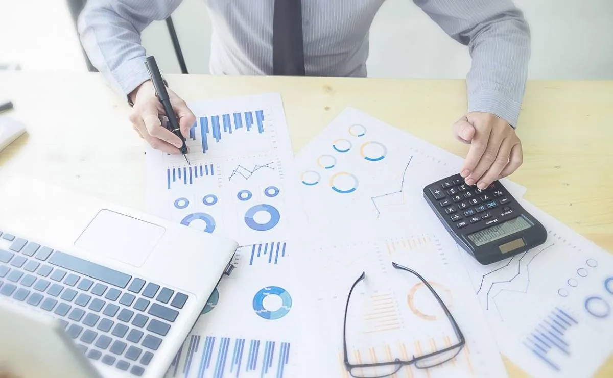 Persona analizando documentos financieros con gráficos y datos, usando una calculadora y un portátil en un escritorio de madera.