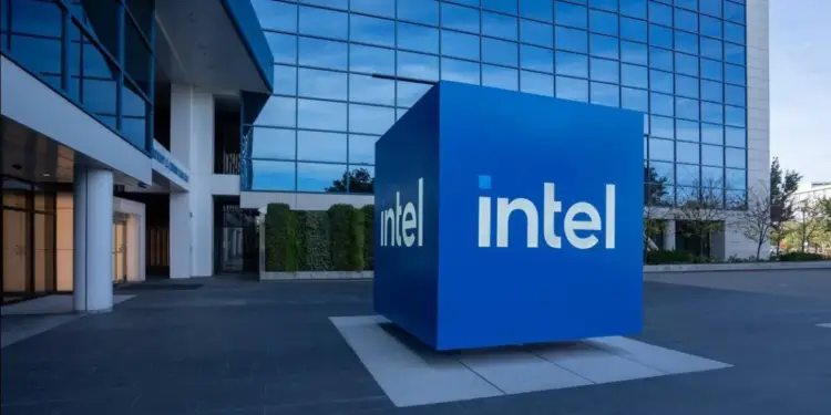 Cubo azul con el logo 'intel' frente a un moderno edificio de oficinas con fachada de cristal azul. Simboliza la infraestructura de una empresa tecnológica global.