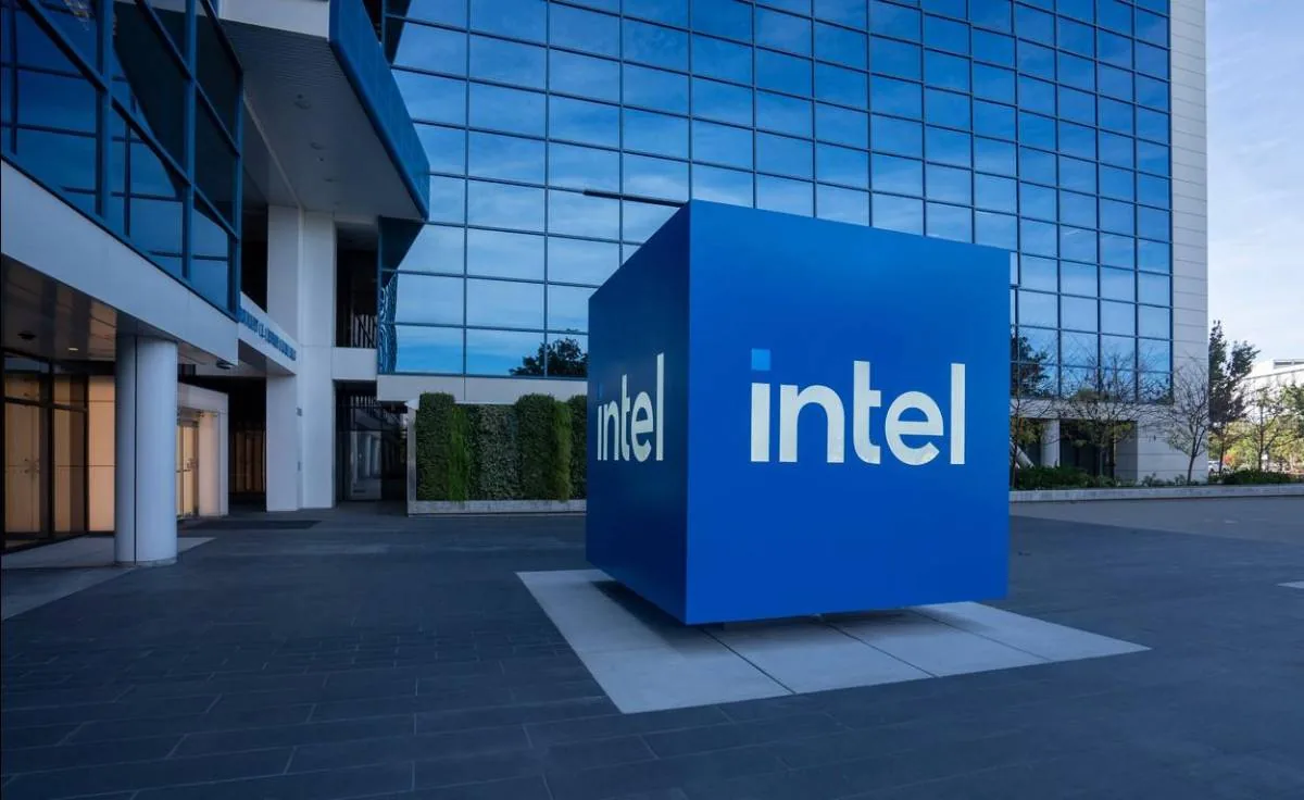 Cubo azul con el logo 'intel' frente a un moderno edificio de oficinas con fachada de cristal azul. Simboliza la infraestructura de una empresa tecnológica global.