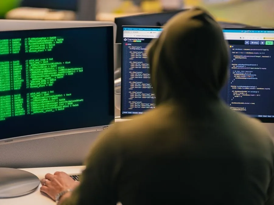 Persona con capucha trabajando frente a múltiples pantallas con líneas de código y simuladores de programación.