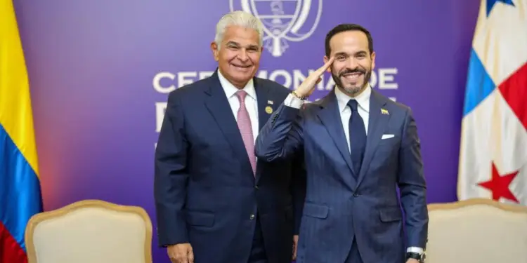 Dos líderes en traje oscuro sonríen en un evento oficial. Uno saluda militarmente. Al fondo, banderas de Colombia y Panamá y un emblema.
