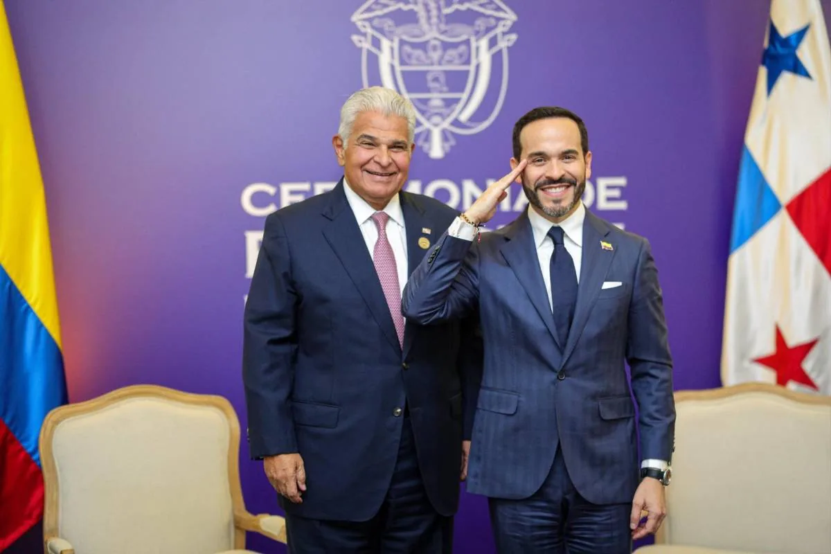 Dos líderes en traje oscuro sonríen en un evento oficial. Uno saluda militarmente. Al fondo, banderas de Colombia y Panamá y un emblema.
