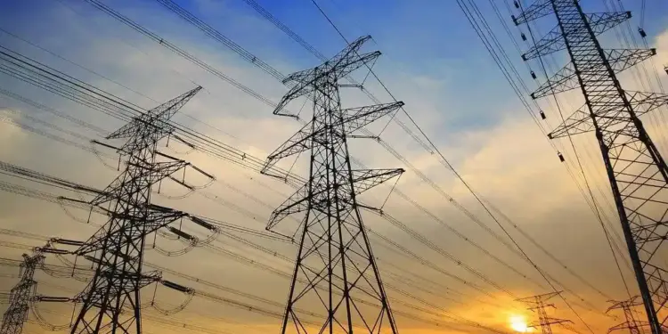 Múltiples torres de alta tensión con cables eléctricos cruzando un cielo nublado al atardecer, con tonos naranjas y azules.