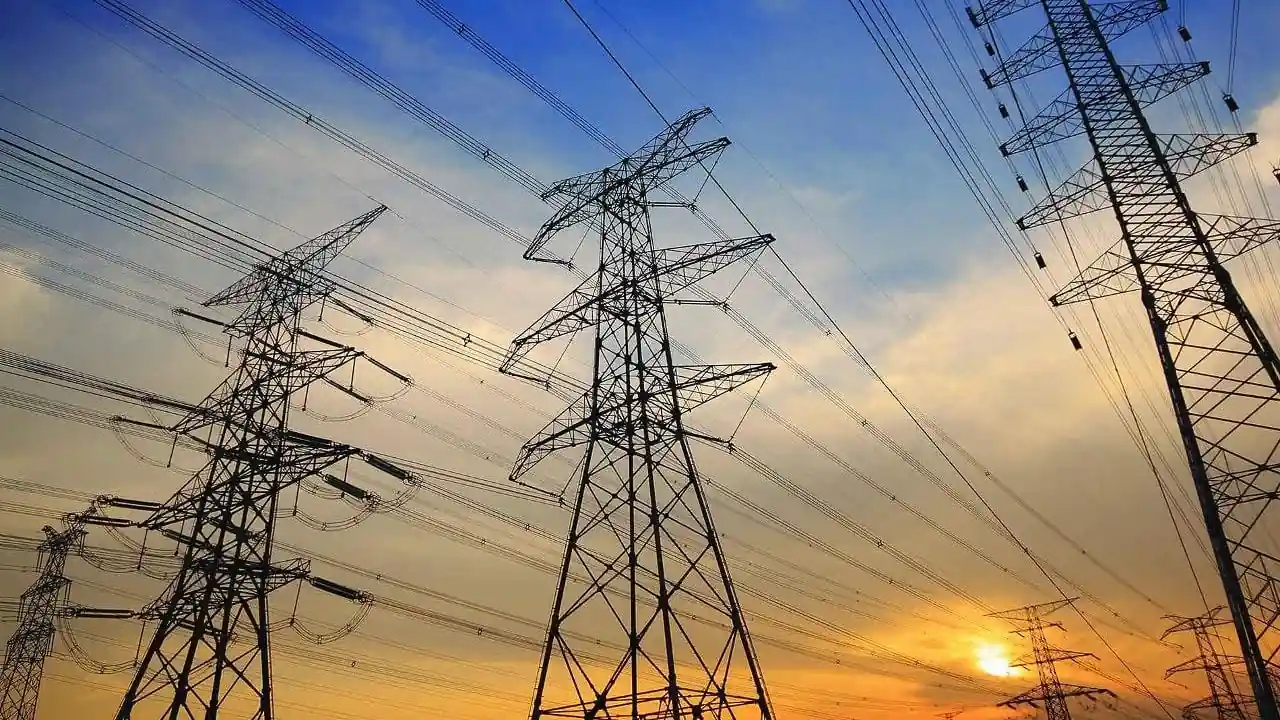 Múltiples torres de alta tensión con cables eléctricos cruzando un cielo nublado al atardecer, con tonos naranjas y azules.