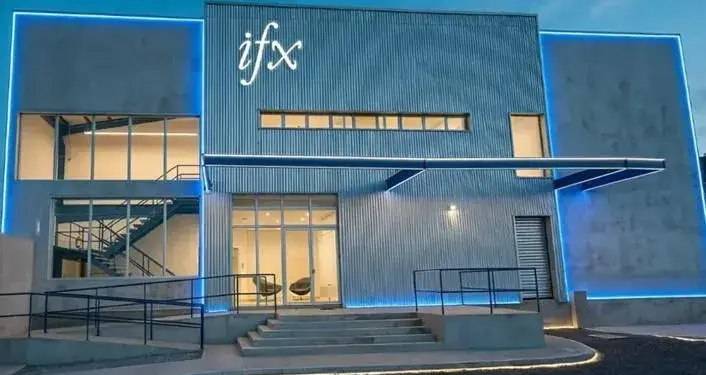 Vista exterior de un edificio moderno de dos pisos con el letrero 'ifx' iluminado. Presenta fachada de hormigón y metal corrugado, grandes ventanales, una escalera interior y luces LED azules que resaltan su diseño. Incluye rampa de acceso.