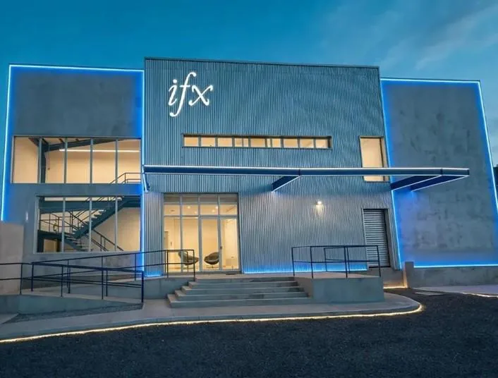 Vista exterior de un edificio moderno de dos pisos con el letrero 'ifx' iluminado. Presenta fachada de hormigón y metal corrugado, grandes ventanales, una escalera interior y luces LED azules que resaltan su diseño. Incluye rampa de acceso.