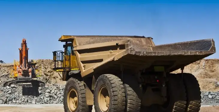 Un gran camión volquete de color ocre en primer plano y una excavadora naranja en un sitio de extracción de recursos, bajo un cielo azul.