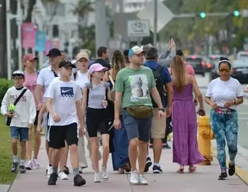 Grupo diverso de personas, incluyendo adultos y niños, caminando por una acera concurrida en un día soleado. Al fondo se ven edificios, vehículos y señales de tráfico, sugiriendo una zona urbana activa.