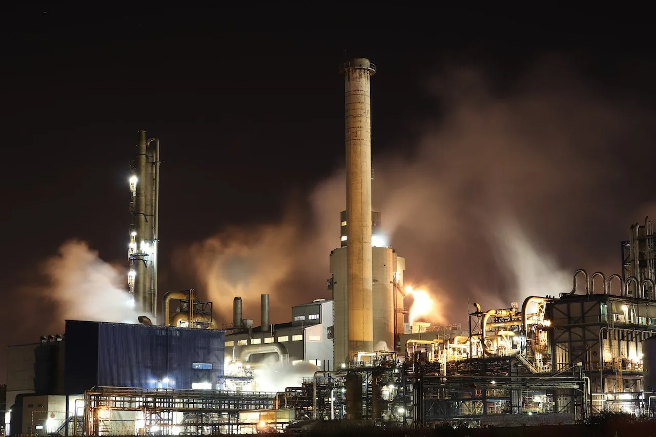 Vista nocturna de un complejo industrial con grandes chimeneas, tuberías y edificios. Se observa humo y vapor saliendo, iluminado por luces intensas y destellos de fuego, indicando actividad productiva.