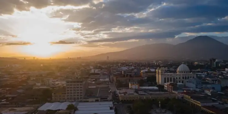 Vista aérea de una ciudad al atardecer, con el sol bajo en el horizonte, edificios urbanos, calles y montañas al fondo bajo un cielo nublado.