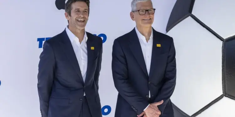 Dos ejecutivos sonrientes en trajes oscuros y camisas blancas, con pines amarillos. Detrás, logo de Apple y un balón de fútbol gigante.