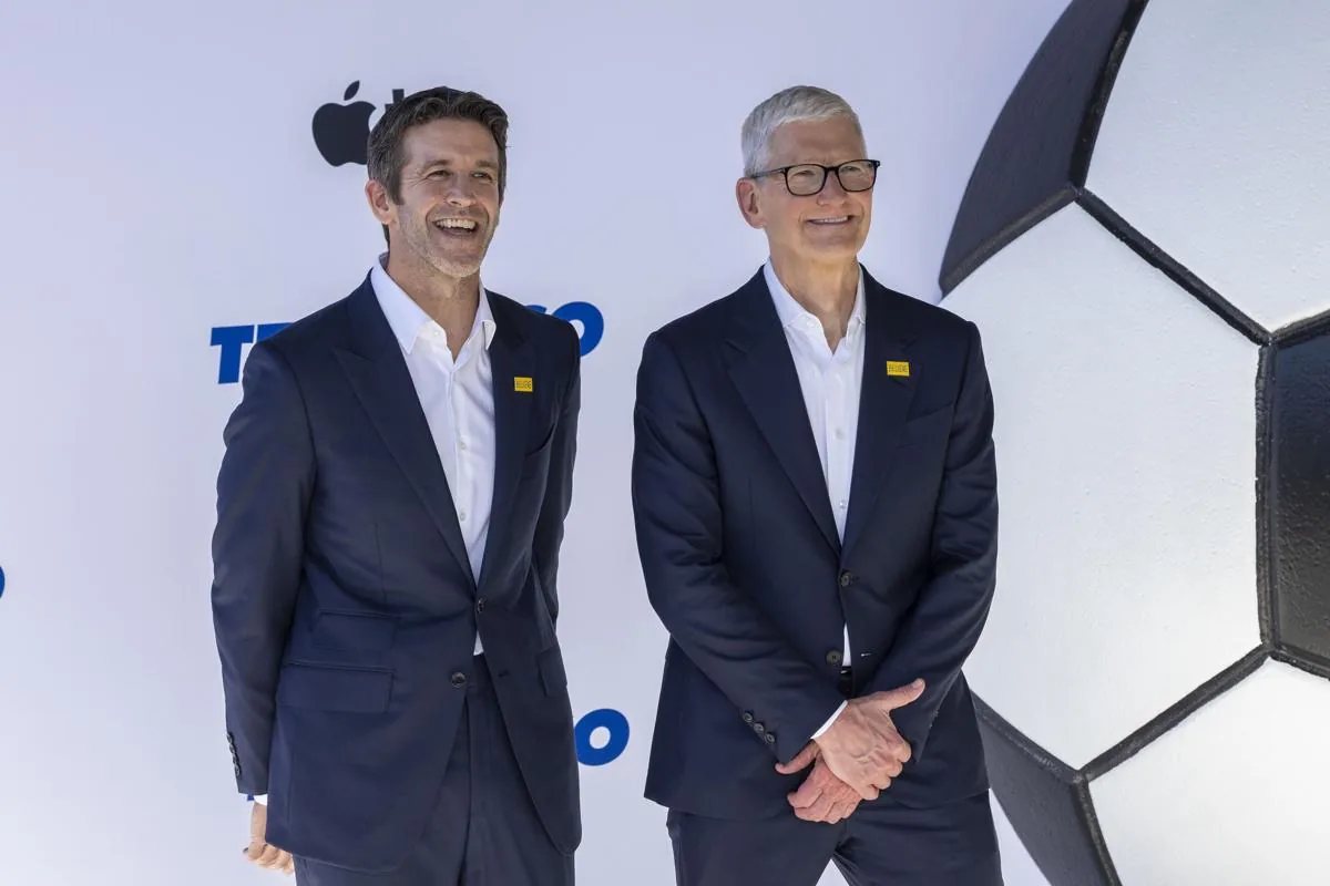 Dos ejecutivos sonrientes en trajes oscuros y camisas blancas, con pines amarillos. Detrás, logo de Apple y un balón de fútbol gigante.