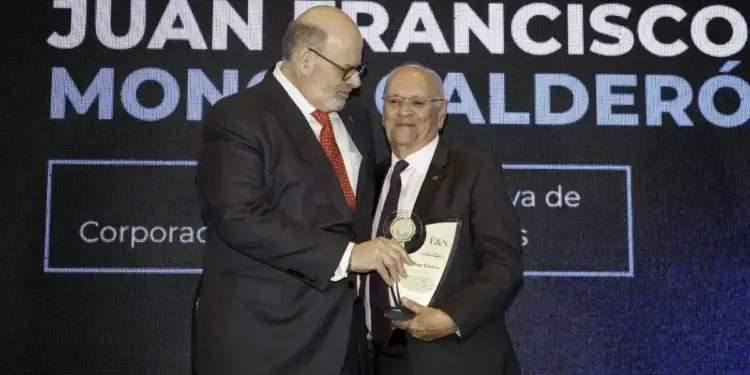 Dos hombres con traje formal compartiendo un momento de entrega de un reconocimiento en un evento con pantalla al fondo.