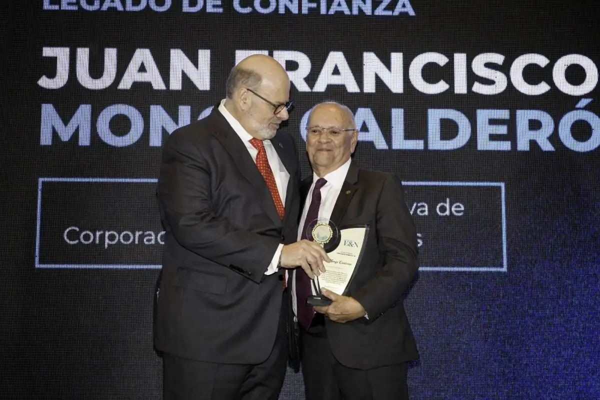 Dos hombres con traje formal compartiendo un momento de entrega de un reconocimiento en un evento con pantalla al fondo.