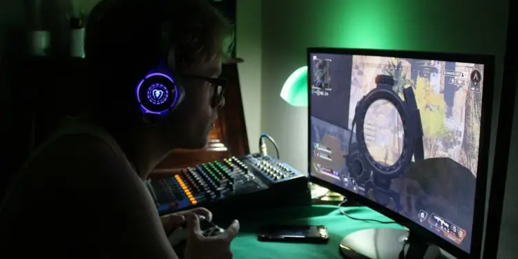 Joven con gafas y auriculares luminosos jugando a videojuegos en un monitor frente a una mesa de mezclas de audio.