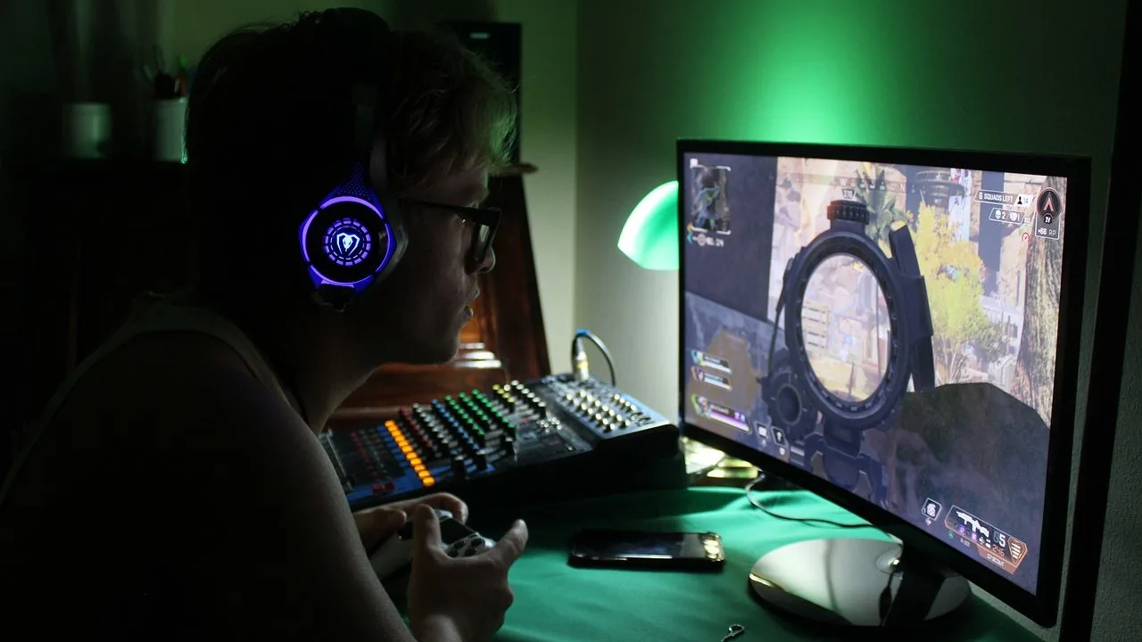 Joven con gafas y auriculares luminosos jugando a videojuegos en un monitor frente a una mesa de mezclas de audio.