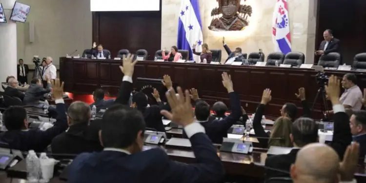 Vista de una asamblea o congreso con múltiples personas levantando sus manos para votar. Banderas nacionales y emblemas visibles.