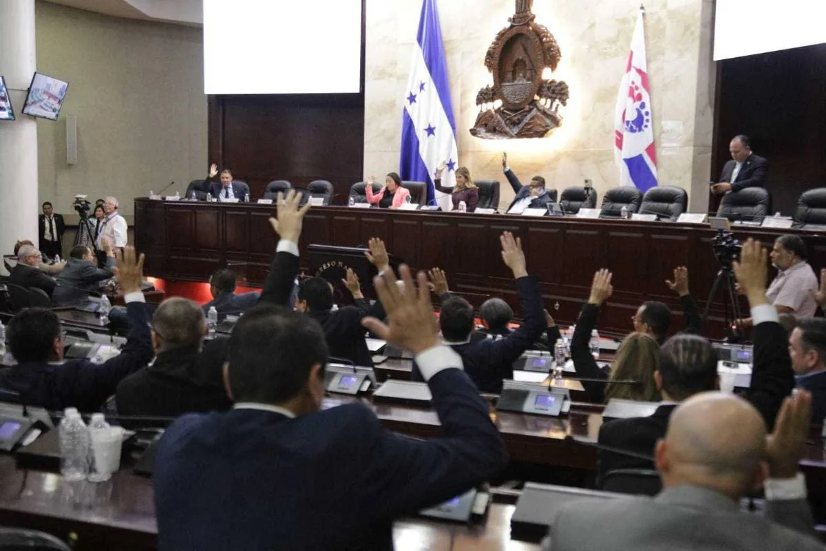Vista de una asamblea o congreso con múltiples personas levantando sus manos para votar. Banderas nacionales y emblemas visibles.