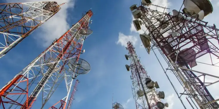Múltiples torres de telecomunicaciones de celosía metálica, pintadas de rojo y blanco, con antenas parabólicas y paneles, contra un cielo azul con nubes. Representa la infraestructura de comunicación.