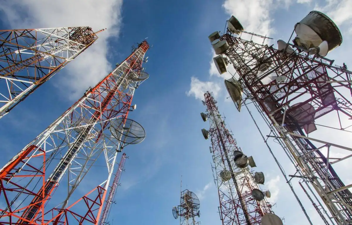 Múltiples torres de telecomunicaciones de celosía metálica, pintadas de rojo y blanco, con antenas parabólicas y paneles, contra un cielo azul con nubes. Representa la infraestructura de comunicación.
