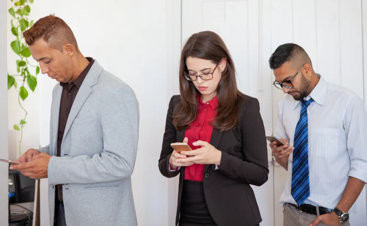 Tres profesionales de pie mirando fijamente las pantallas de sus teléfonos inteligentes en una oficina.