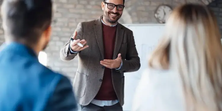 Hombre sonriente con gafas y blazer marrón explicando conceptos en una reunión de negocios, con un rotulador en mano, frente a dos asistentes.