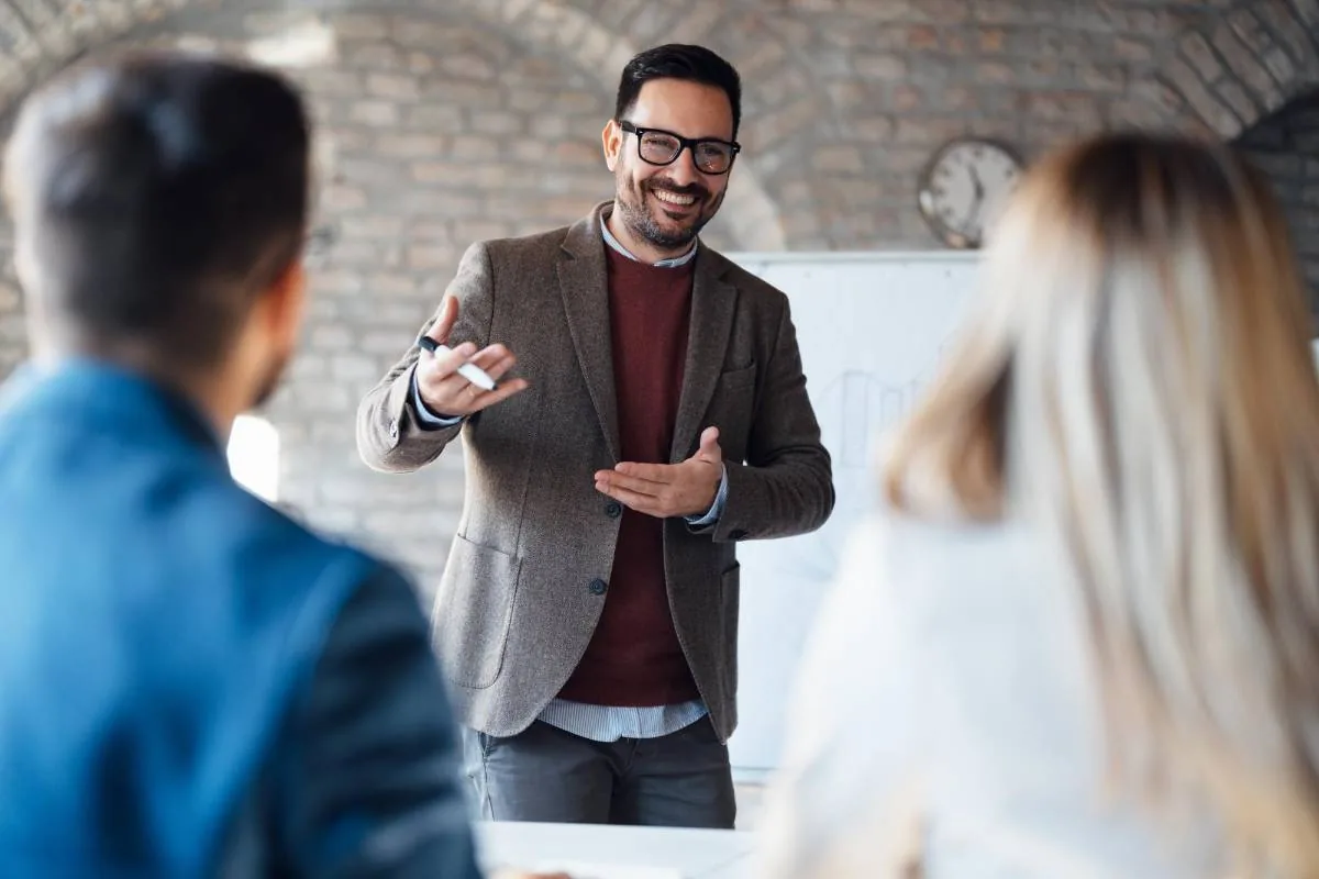 Hombre sonriente con gafas y blazer marrón explicando conceptos en una reunión de negocios, con un rotulador en mano, frente a dos asistentes.