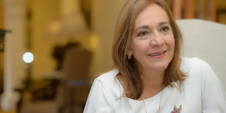 Retrato de mujer profesional de mediana edad con blusa blanca y broche de colibrí. Sonríe, fondo interior difuminado.