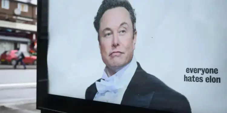 Un cartel publicitario muestra a Elon Musk con esmoquin y pajarita blanca, con la frase "everyone hates elon". Al fondo, una calle borrosa con un coche rojo y una persona.