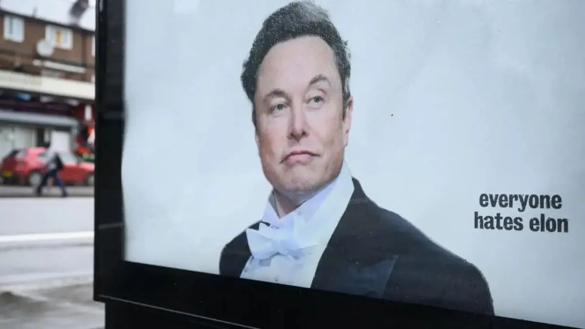 Un cartel publicitario muestra a Elon Musk con esmoquin y pajarita blanca, con la frase "everyone hates elon". Al fondo, una calle borrosa con un coche rojo y una persona.