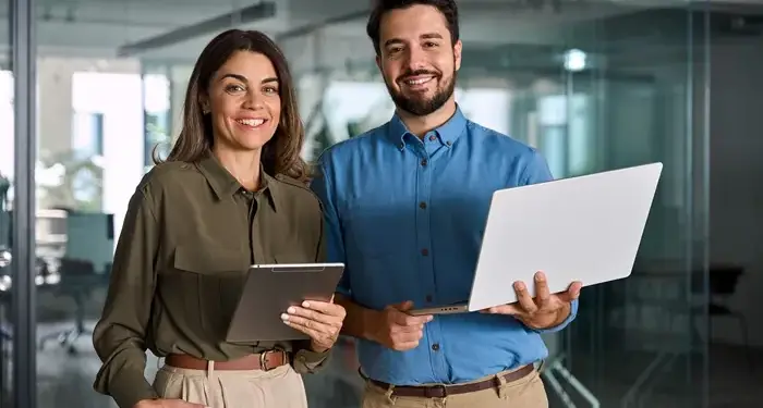 Hombre y mujer profesionales sonrientes sosteniendo una laptop y una tableta en una oficina moderna.