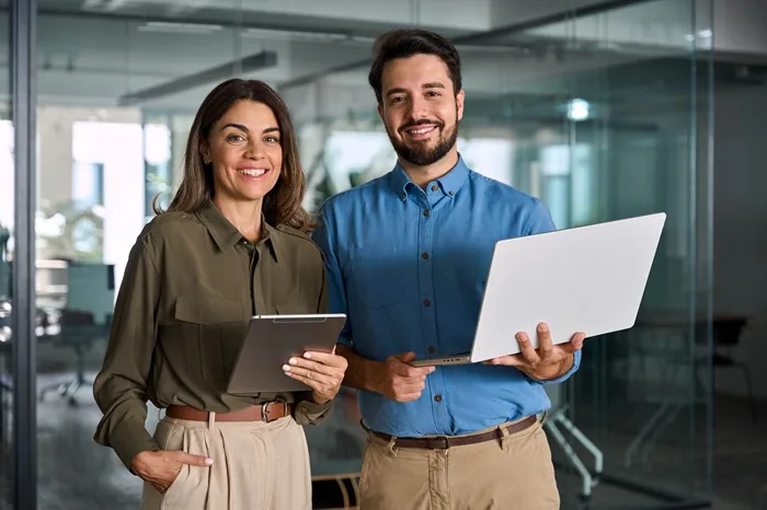 Hombre y mujer profesionales sonrientes sosteniendo una laptop y una tableta en una oficina moderna.