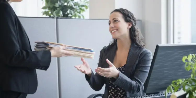 Dos profesionales en una oficina intercambian documentos. Una mujer entrega una pila de archivos a otra sentada en un escritorio con un ordenador, sonriendo.