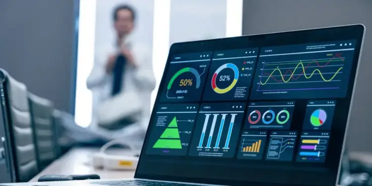Laptop mostrando un dashboard con gráficos de análisis de datos financieros y empresariales. Una persona desenfocada en el fondo.