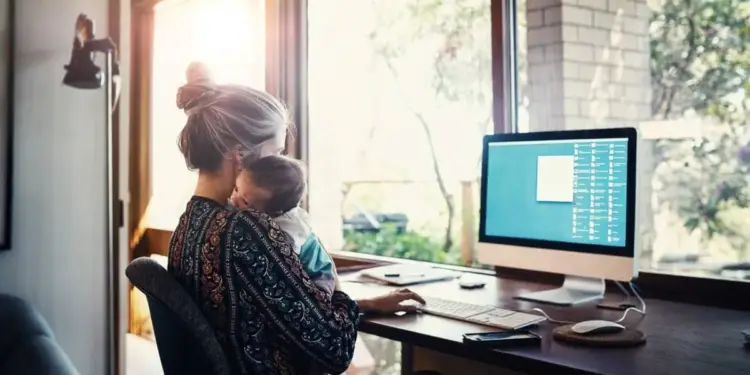 Mujer con bebé en brazos trabajando en un escritorio con computadora frente a una ventana. Representa el teletrabajo y la conciliación familiar.