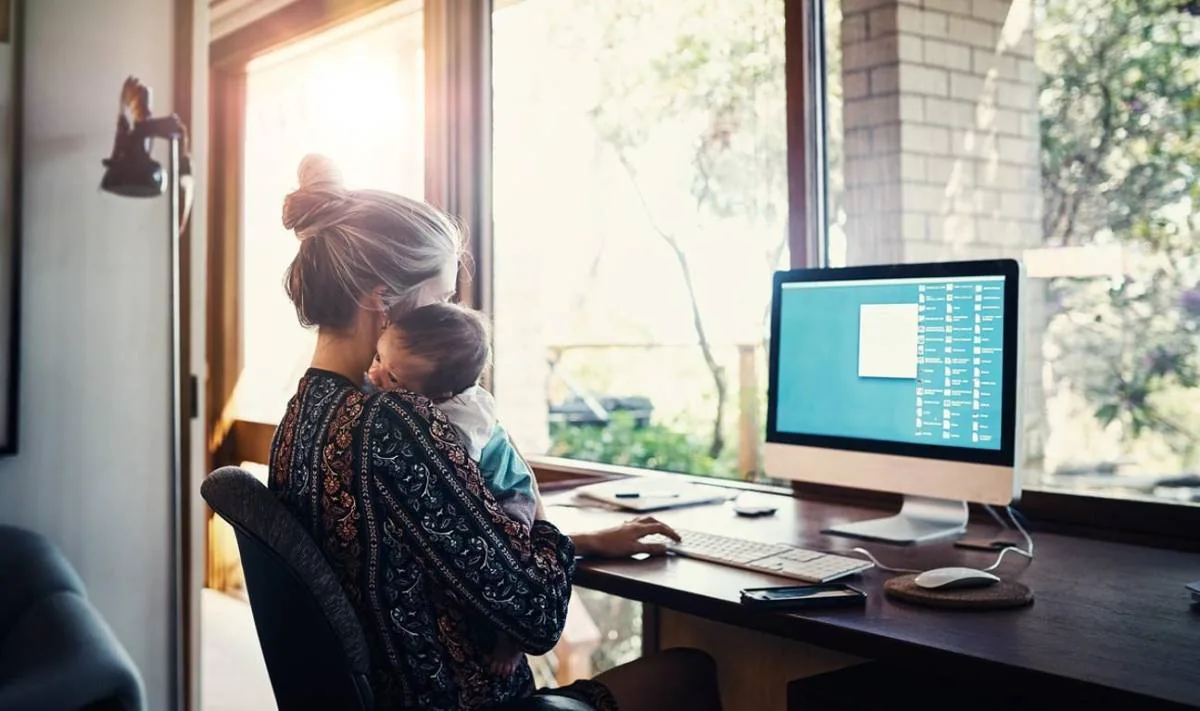 Mujer con bebé en brazos trabajando en un escritorio con computadora frente a una ventana. Representa el teletrabajo y la conciliación familiar.
