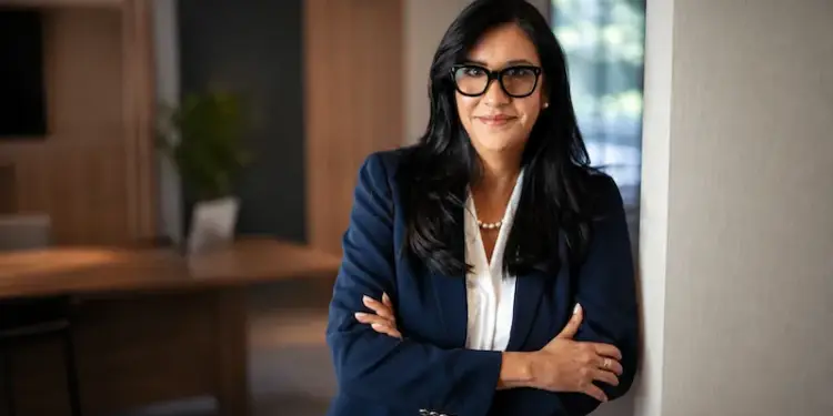 Mujer profesional sonriente con gafas y blazer azul, brazos cruzados, en un entorno de oficina moderno. Representa éxito empresarial.