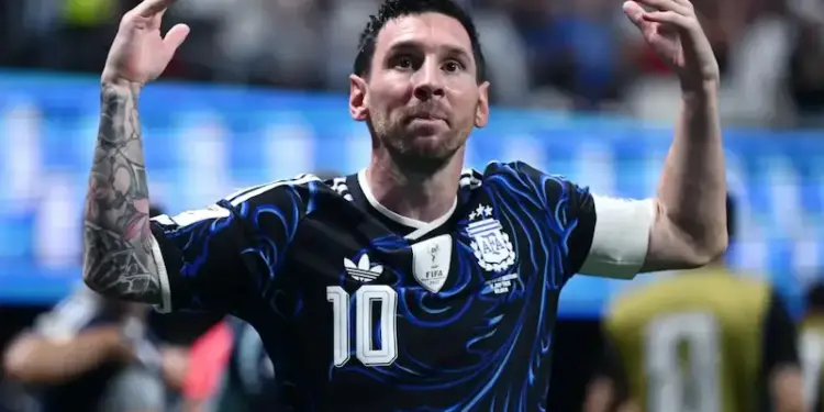 Lionel Messi vistiendo una camiseta oscura con detalles azules y el número 10, celebrando con las manos levantadas en un estadio.