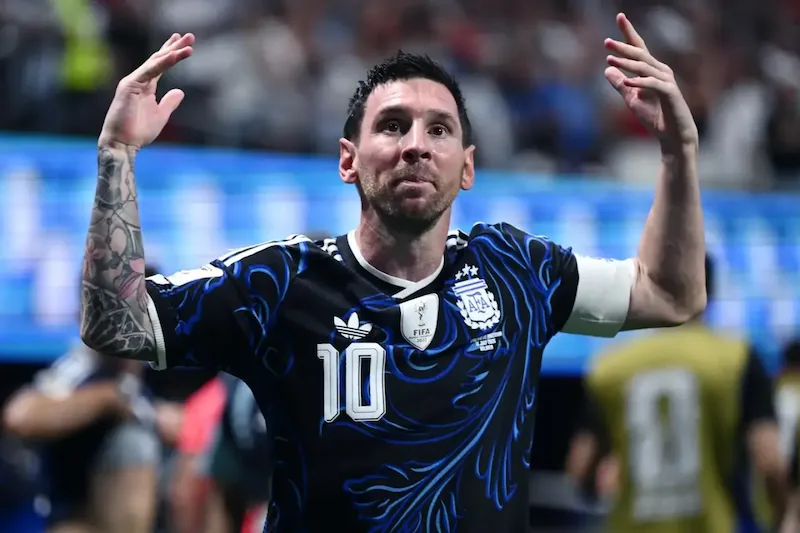 Lionel Messi vistiendo una camiseta oscura con detalles azules y el número 10, celebrando con las manos levantadas en un estadio.